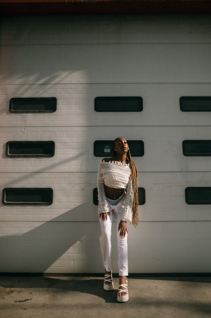 Young woman in a stylish white outfit posing in front of a garage door, bathed in summer sunlight.