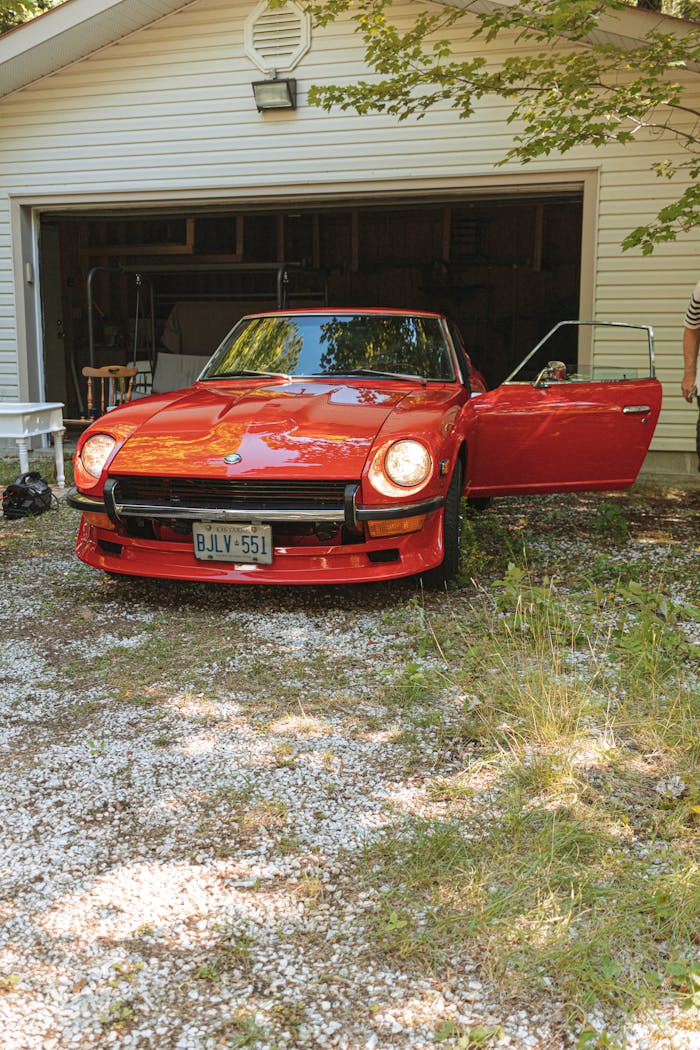Classic red Datsun 240Z with open door parked in a driveway near a garage.